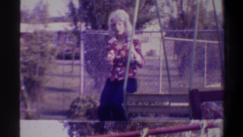 Young blonde woman in floral blouse standing by a backyard swing set; suburban Gainesville, Florida setting circa 1974; 8mm footage with characteristic purple-magenta film shift.
