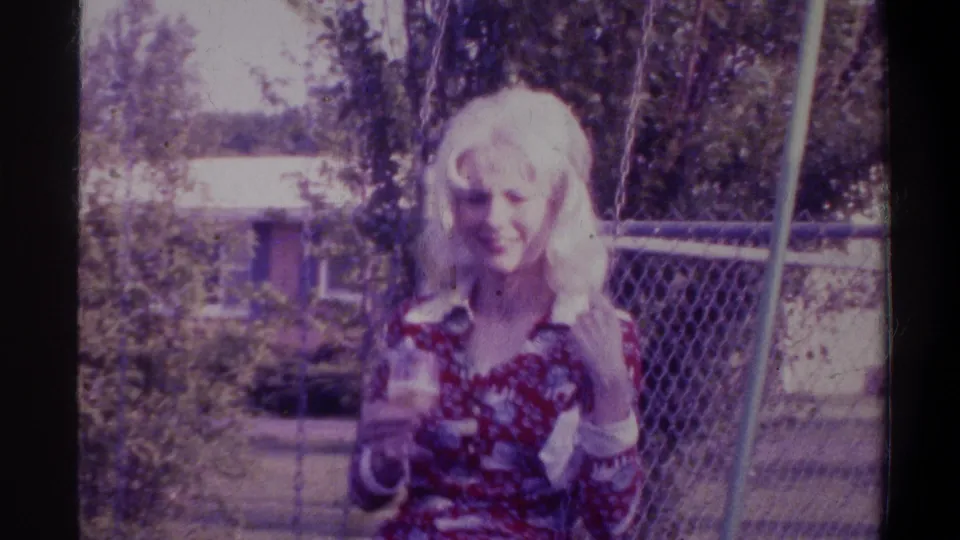 Young blonde woman smiling and holding a drink in a suburban backyard, circa 1974 Gainesville, Florida; red floral blouse visible; 8mm film with purple color cast and grain.