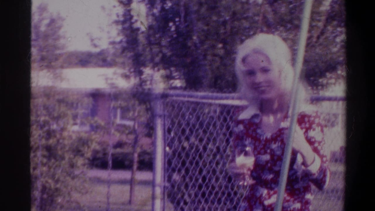Blonde woman in a dark floral dress on a backyard swing, chain-link fence and a low residential building behind her, circa-1974 Gainesville, Florida home movie.