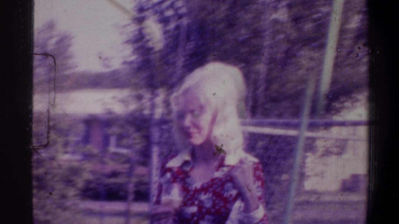 Blonde woman in a floral dress mid-swing with motion blur, chain-link fence and trees behind her, heavy violet color cast on aged 8mm film.