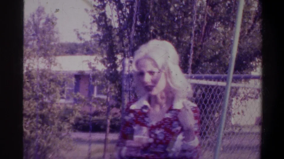 Blonde woman looking downward while possibly holding a glass, backyard with chain-link fence, from a circa-1974 Gainesville, Florida home movie.