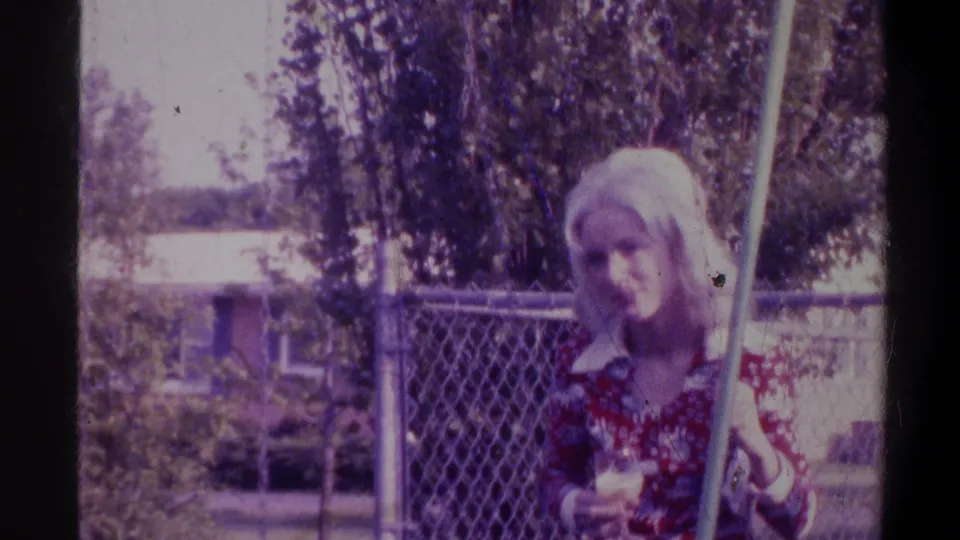 Blonde woman on a backyard swing looking downward, residential neighborhood visible through chain-link fence, Gainesville, Florida, circa 1974.