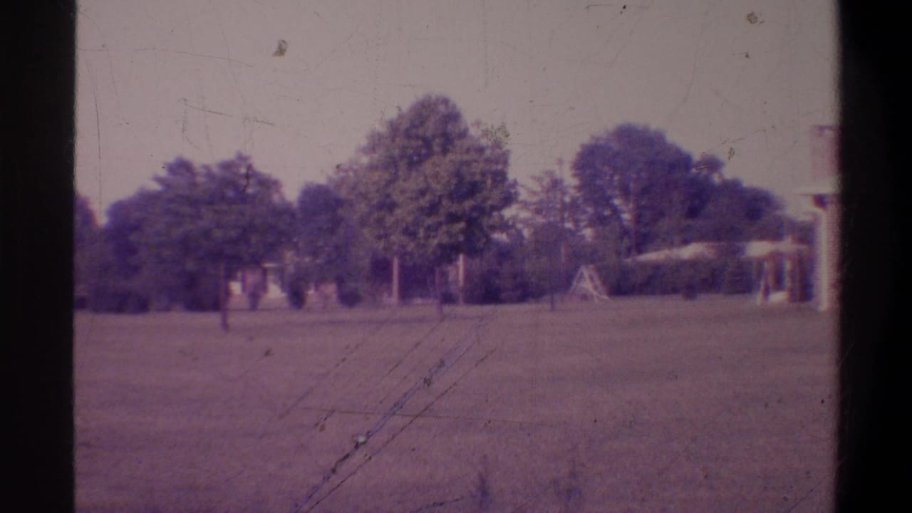 Wide grassy lawn with large trees and a partially visible house, shot on faded 8mm film with a purple color cast.