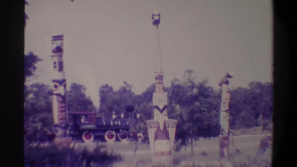 Dark miniature park train locomotive with red wheel detail passing painted totem-style columns in a Florida park, 1974, 8mm film