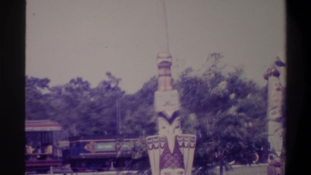 Two elaborately painted totem poles in the foreground with a tourist train entering at left, at what appears to be a Florida family attraction, c. 1974.