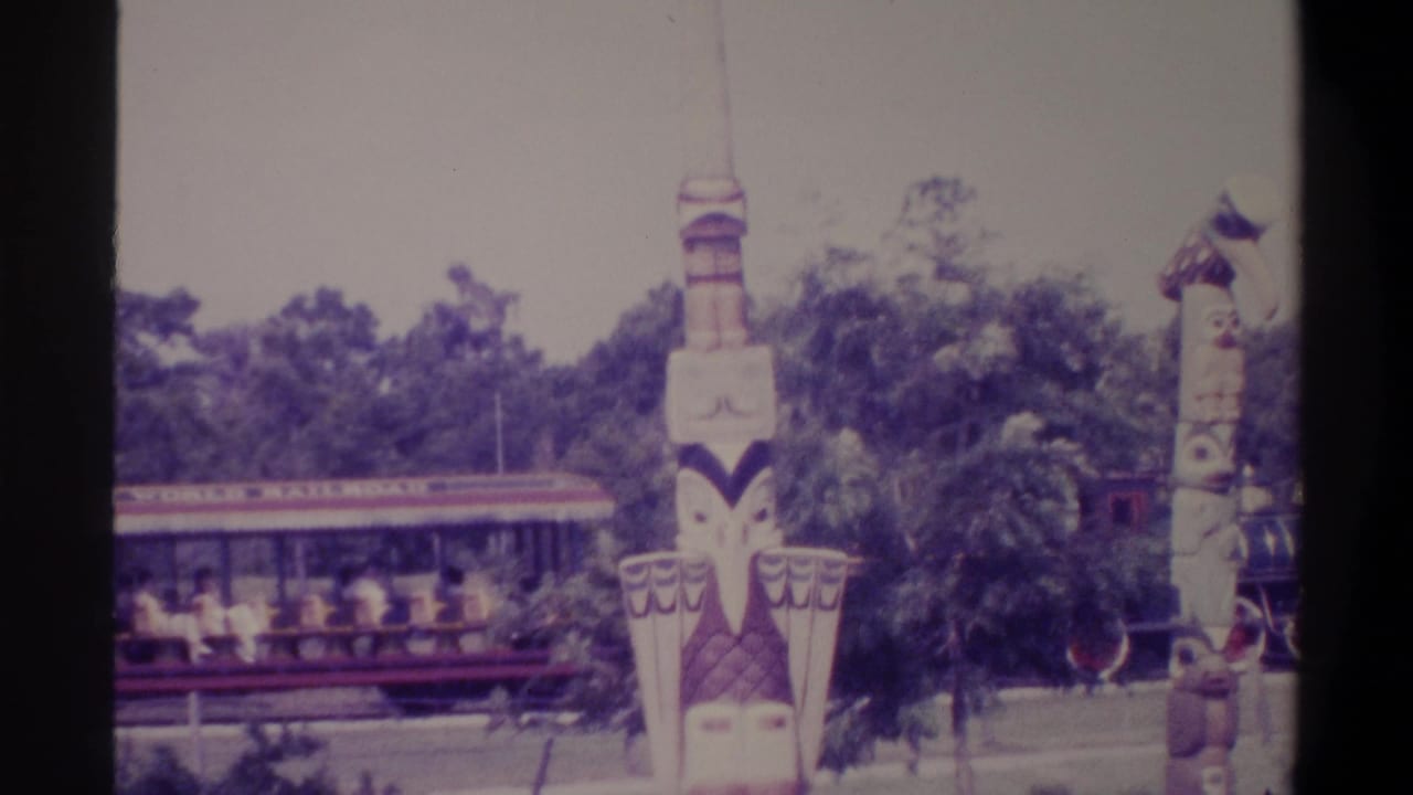 Adult passengers visible in open-air tourist train cars moving past painted totem poles, likely at a Florida family attraction, c. 1974.