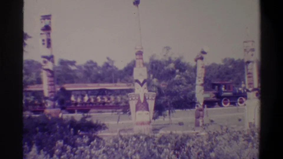 Several painted totem poles at what appears to be a Florida tourist attraction, with a small locomotive and open train cars visible behind them, c. 1974.