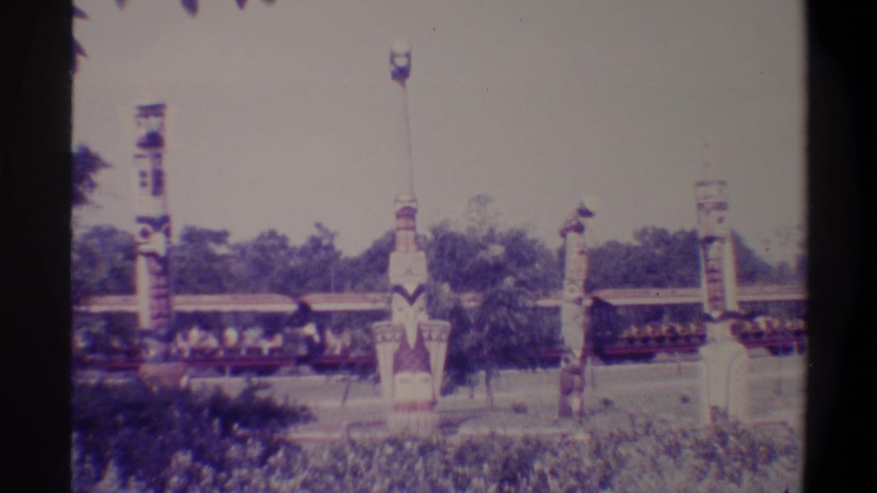 Visitor-shot 8mm frame from a Florida tourist attraction, c. 1974, showing painted totem poles and a passing open-air ride train with passengers.