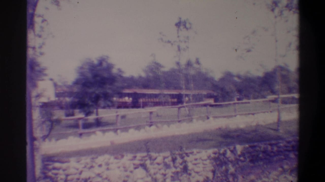 Train or platform structure at left, stone-edged track border and wooden rail fence in foreground, trees behind, faded 8mm color.