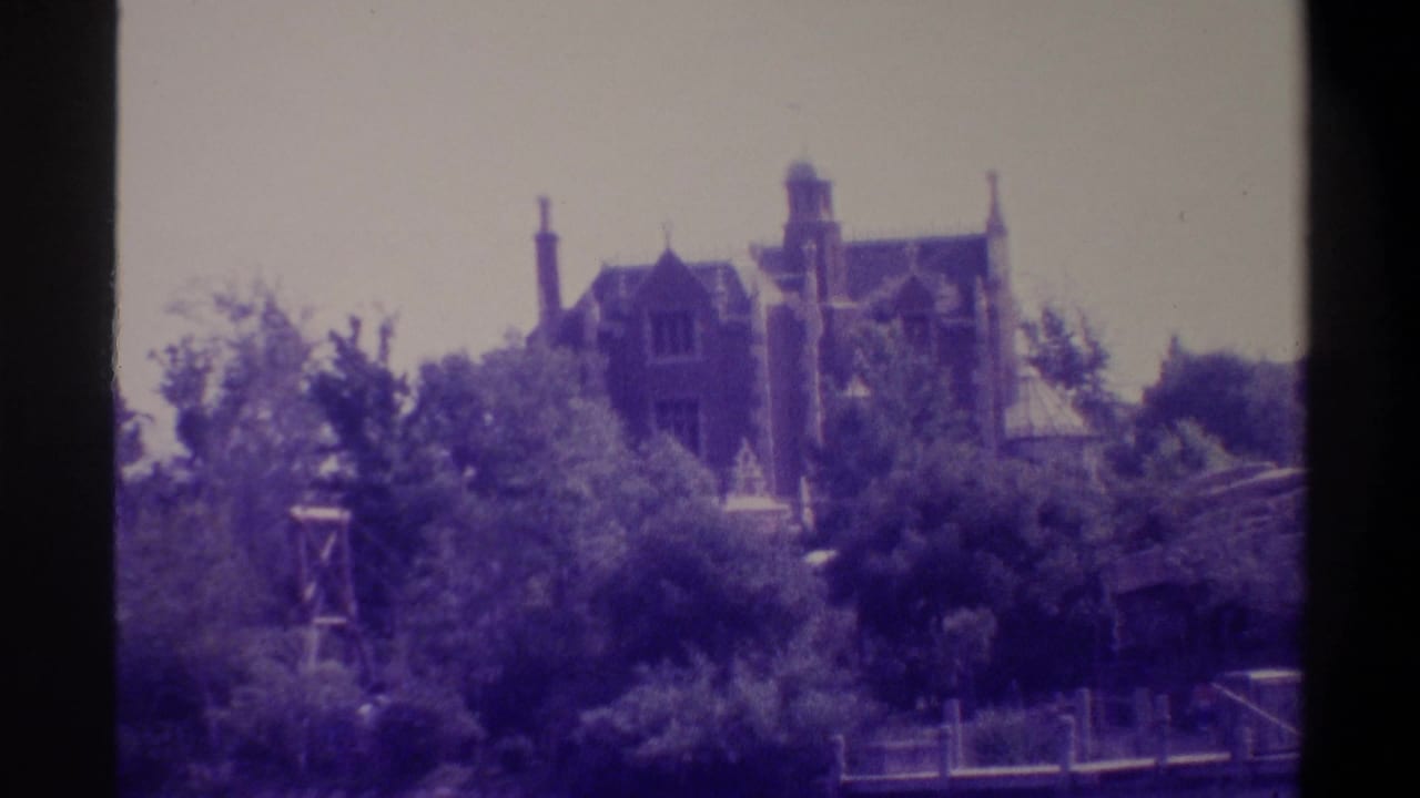 Gothic Revival mansion with pointed towers and chimneys rising above a dense treeline, metal framework structure in foreground.