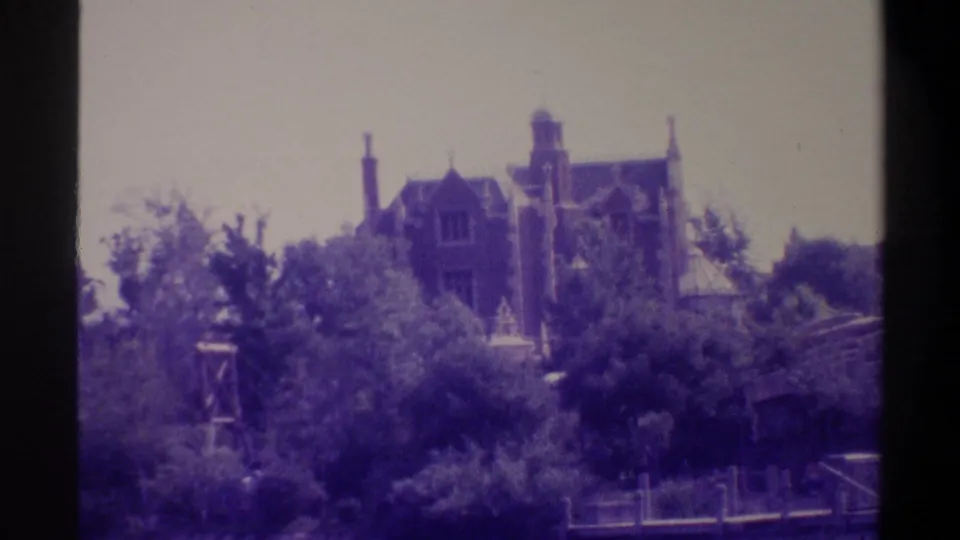 Gothic Revival mansion with pointed towers and brick chimneys visible above subtropical trees, with a metal scaffolding or framework structure in the foreground; 8mm film, Gainesville Florida area, circa 1974.