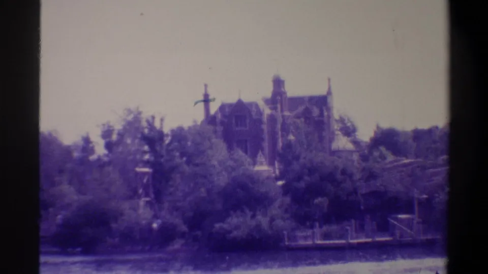 Gothic Revival mansion with multiple towers and chimneys viewed across a calm river or lake, dense subtropical treeline between building and water; 8mm home movie, Gainesville Florida area, circa 1974.