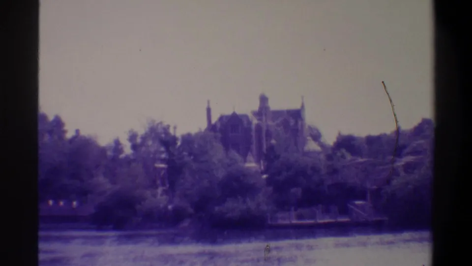 Gothic Revival mansion with towers and chimneys rising above a subtropical treeline, viewed across a calm waterway; 8mm home movie, Gainesville Florida area, circa 1974.