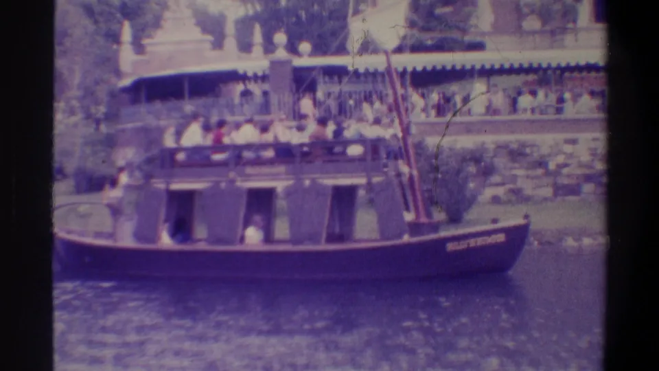 1974 8mm film still of a tourist boat tour on a Florida waterway with passengers aboard on both levels; ornate landmark building visible through subtropical foliage in background.