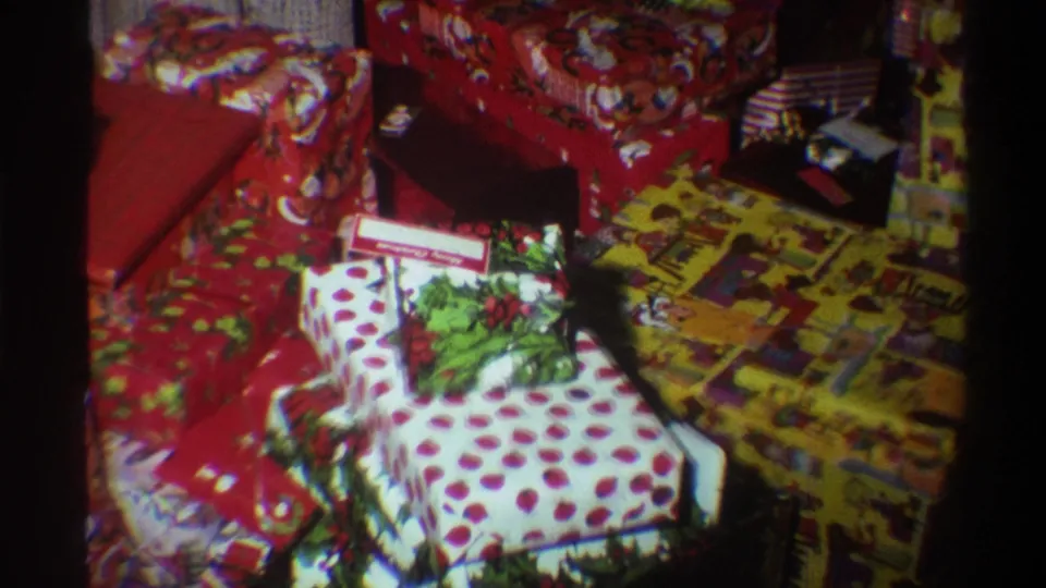 8mm footage circa 1975 panning across Christmas gifts; white polka-dot box with gift tag prominent, yellow multi-pattern paper beneath.