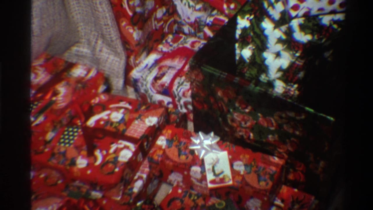 Close-up 8mm home movie frame from Gainesville, Florida, 1975, showing a pile of Christmas-wrapped gifts in Santa and holly print paper with a visible gift tag.