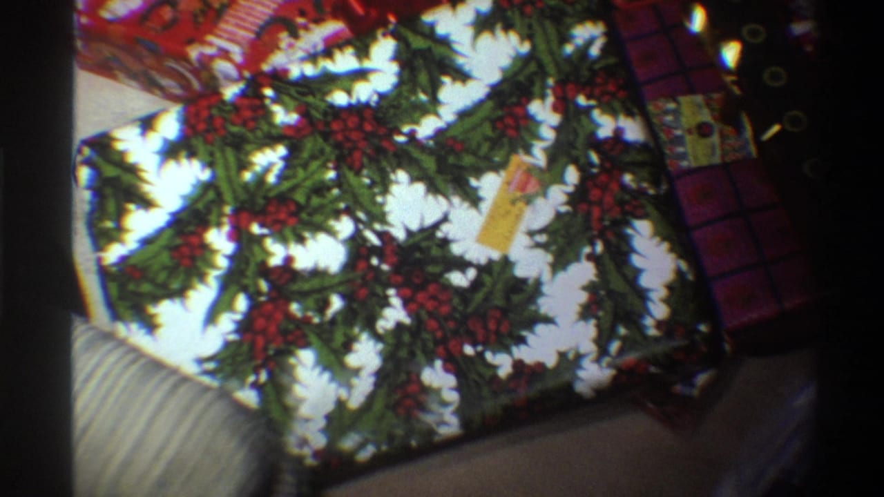 8mm home movie frame from Gainesville, Florida, 1975, showing a large flat present in holly-print paper on a couch with other gifts visible in the background.