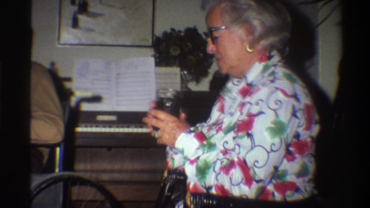 Older woman in floral blouse and glasses holding a glass near a home organ with sheet music, 8mm home movie frame.