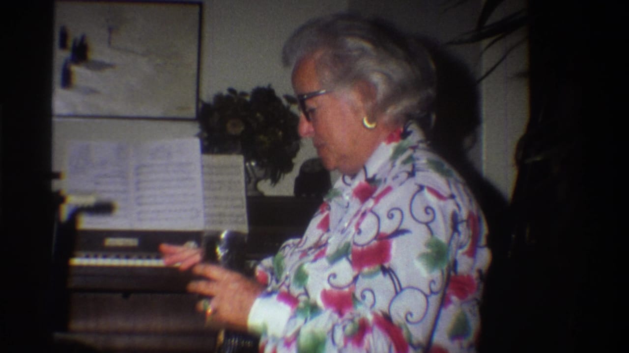 Older woman in 1970s floral blouse in profile at a home organ; sheet music on stand and artificial flower arrangement visible in a private domestic interior, circa 1975.