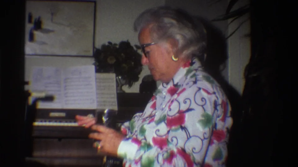 Older woman in 1970s floral blouse in profile at a home organ; sheet music on stand and artificial flower arrangement visible in a private domestic interior, circa 1975.