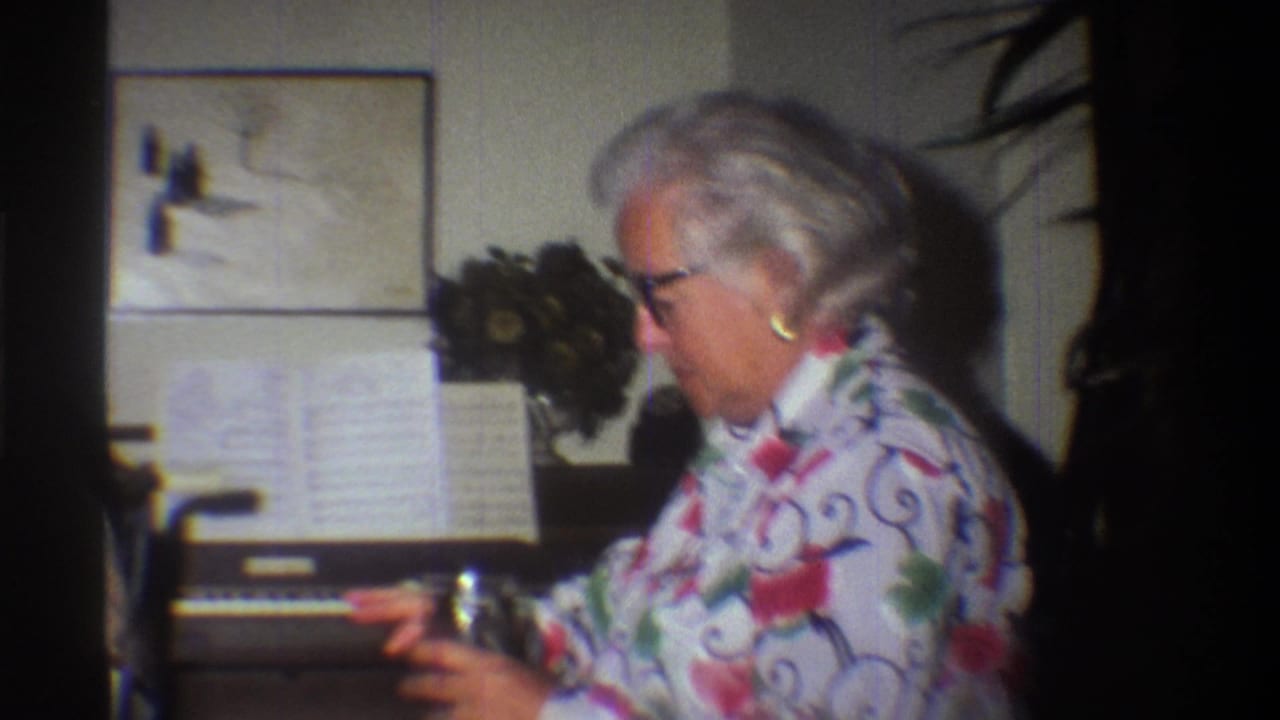 Older woman in 1970s floral blouse at a home organ; warm 8mm color cast and film grain characteristic of mid-1970s amateur film stock, Gainesville Florida circa 1975.