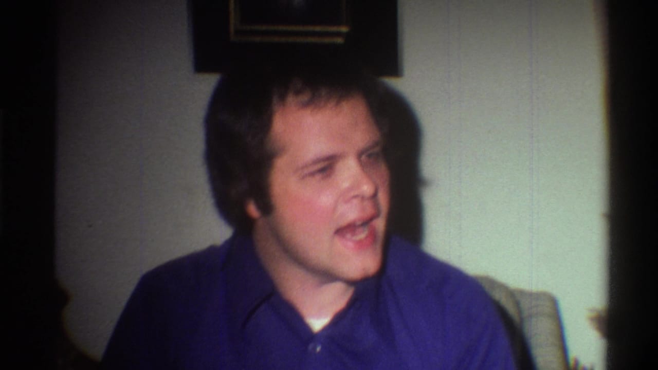 Young adult man in a blue collared polo shirt seated in a domestic living room, caught mid-expression; Gainesville, Florida, circa 1975, 8mm home movie.