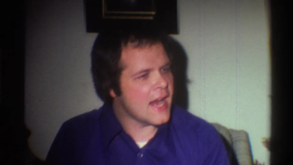Young adult man in a blue collared polo shirt seated in a domestic living room, caught mid-expression; Gainesville, Florida, circa 1975, 8mm home movie.