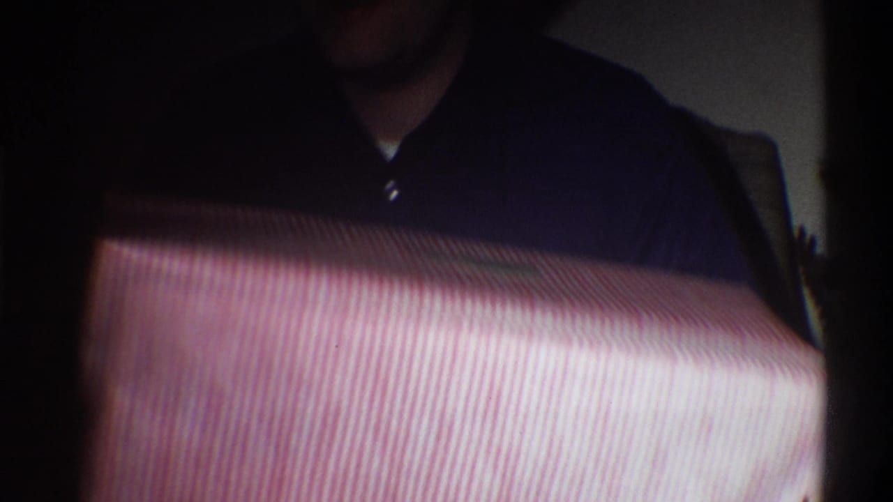 Close-up of a man in a dark shirt holding a large red-and-white striped gift box in a dim indoor setting, circa 1975 Gainesville Florida