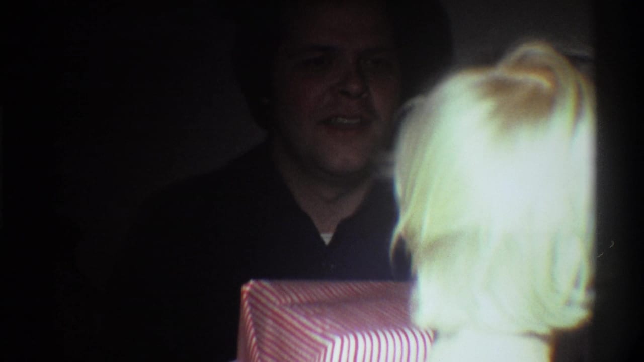 Man with dark hair smiling near a red-and-white striped gift box in a dimly lit room