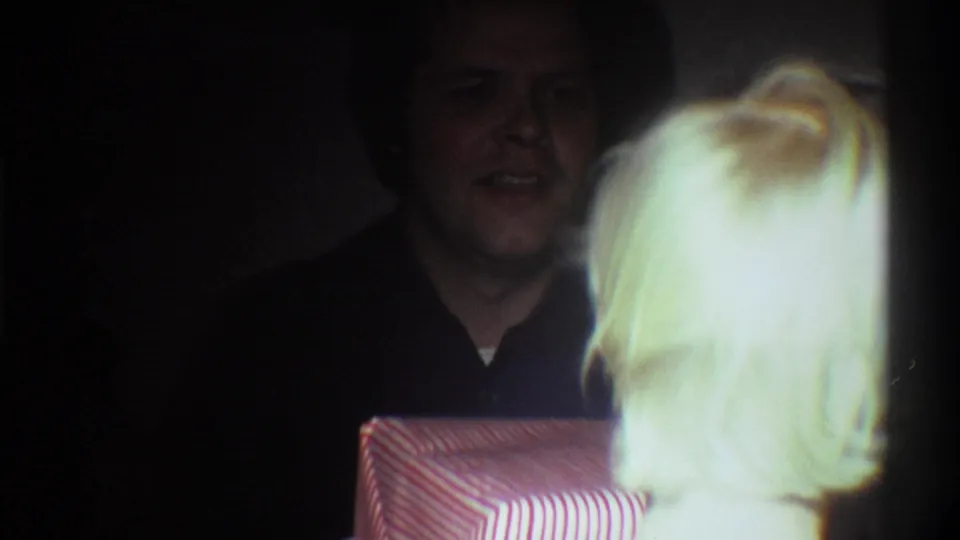 Adult male in dark shirt smiling, with a red-and-white striped gift box visible at lower frame, in a dim domestic interior circa 1975