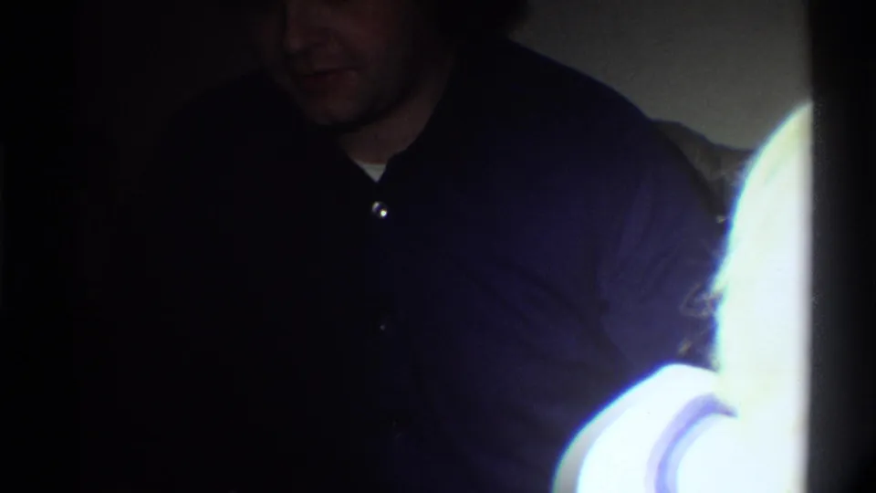 Man in dark shirt bending forward or looking down in a dim home interior, possibly mid-1970s domestic Florida setting