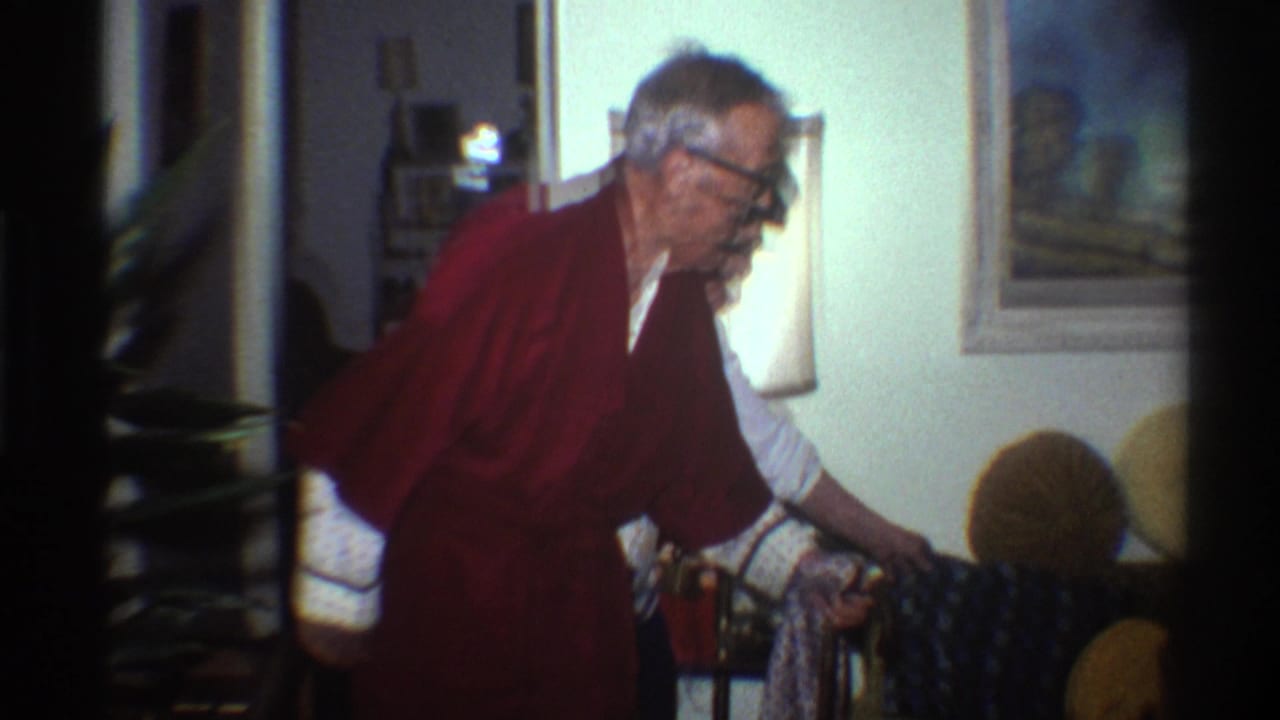 Elderly man in a dark red robe and black-framed glasses leans forward with a cane in a 1970s domestic living room interior, Gainesville, Florida, 1975.