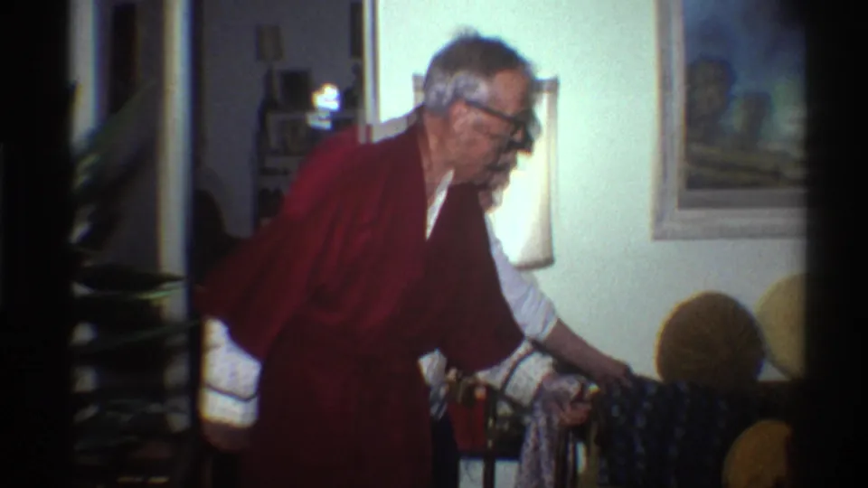 Elderly man in a dark red robe and black-framed glasses leans forward with a cane in a 1970s domestic living room interior, Gainesville, Florida, 1975.
