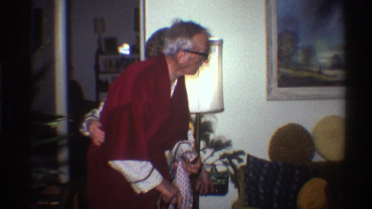 Elderly man in red robe and glasses stands in a living room beside a lit floor lamp and framed landscape painting.