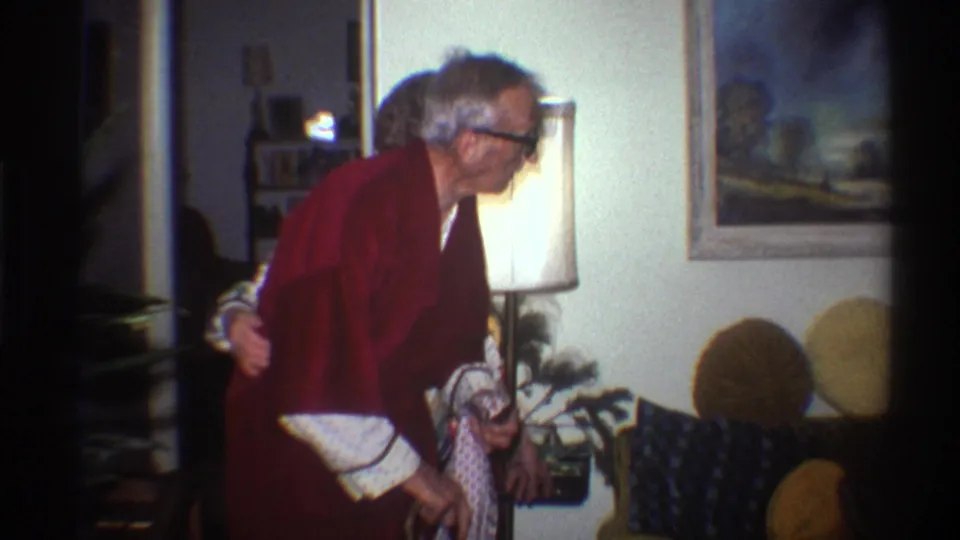 Elderly man in a dark red robe and black-framed glasses stands in a 1970s living room; a floor lamp with white shade and framed landscape oil painting are visible behind him.