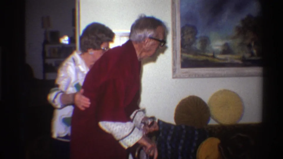 An elderly woman in a white patterned blouse assists an elderly man in a dark red robe in a 1970s domestic living room; a framed landscape painting hangs on the wall behind them.