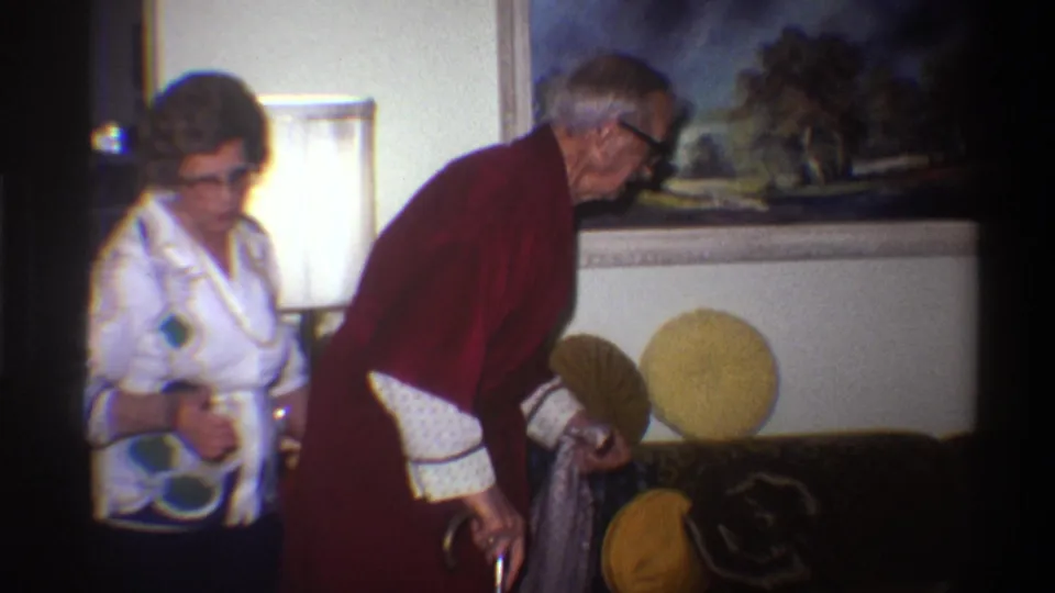 Elderly man in dark red robe gripping a cane and elderly woman in white patterned blouse stand in a 1970s living room; a curtained window and landscape painting are visible behind them.