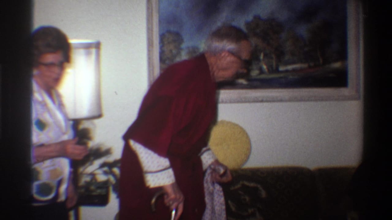 8mm home movie still from 1975: elderly man in dark red robe grips a white cane while leaning forward; a woman in a geometric-print blouse stands to his left in a warmly lit living room.