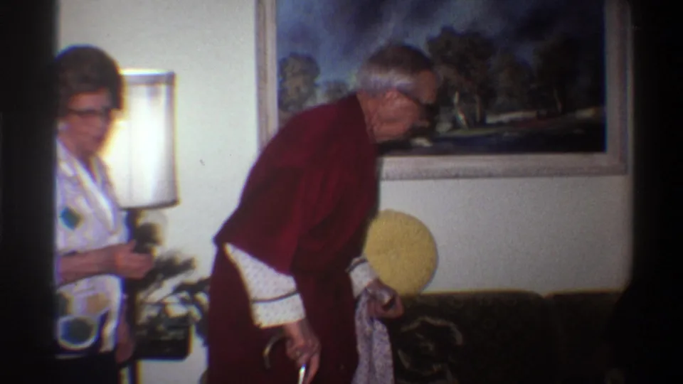 8mm home movie still from 1975: elderly man in dark red robe grips a white cane while leaning forward; a woman in a geometric-print blouse stands to his left in a warmly lit living room.
