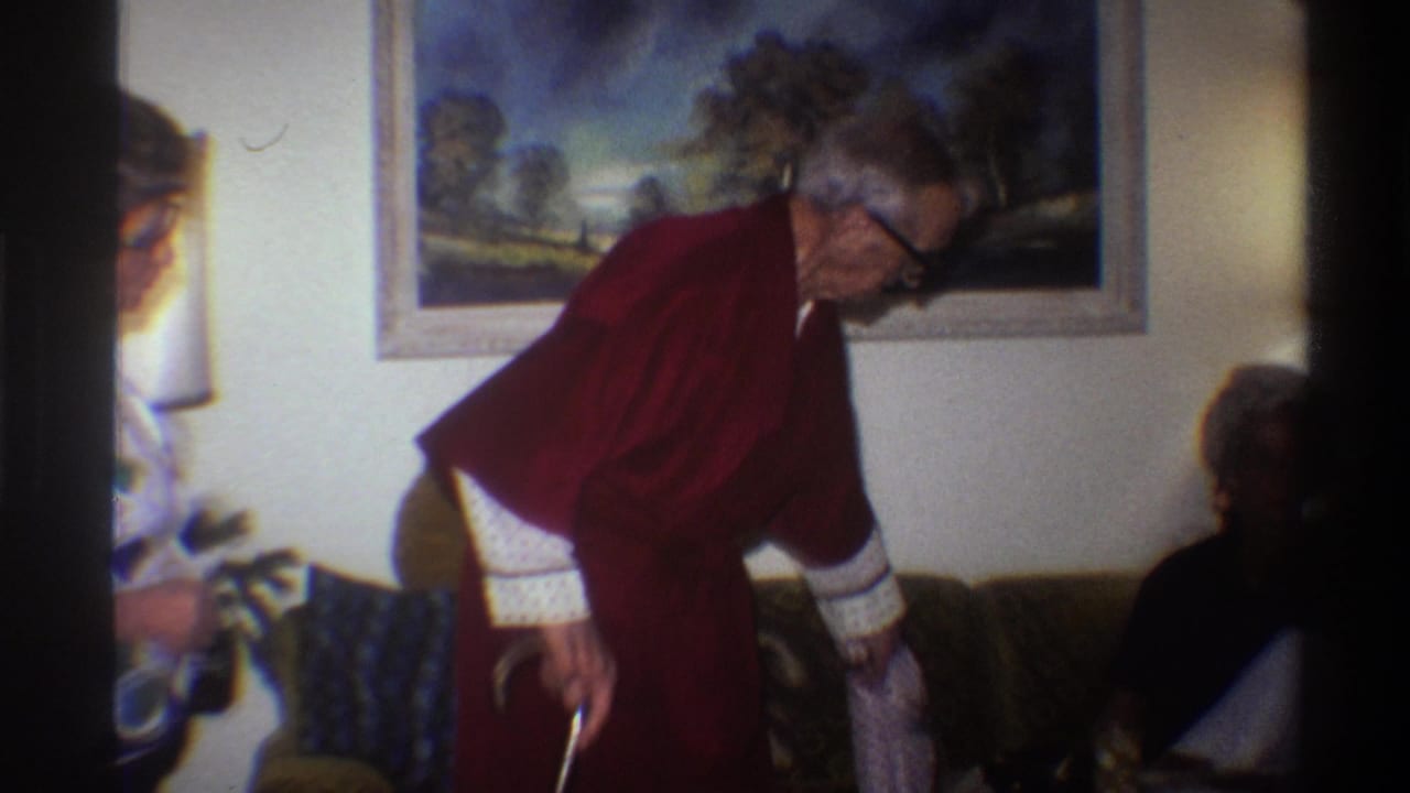 Mid-shot from a 1975 8mm home movie: elderly man in dark red robe with cane bending toward couch; partial silhouette of a second figure visible at right edge; framed landscape painting on wall.