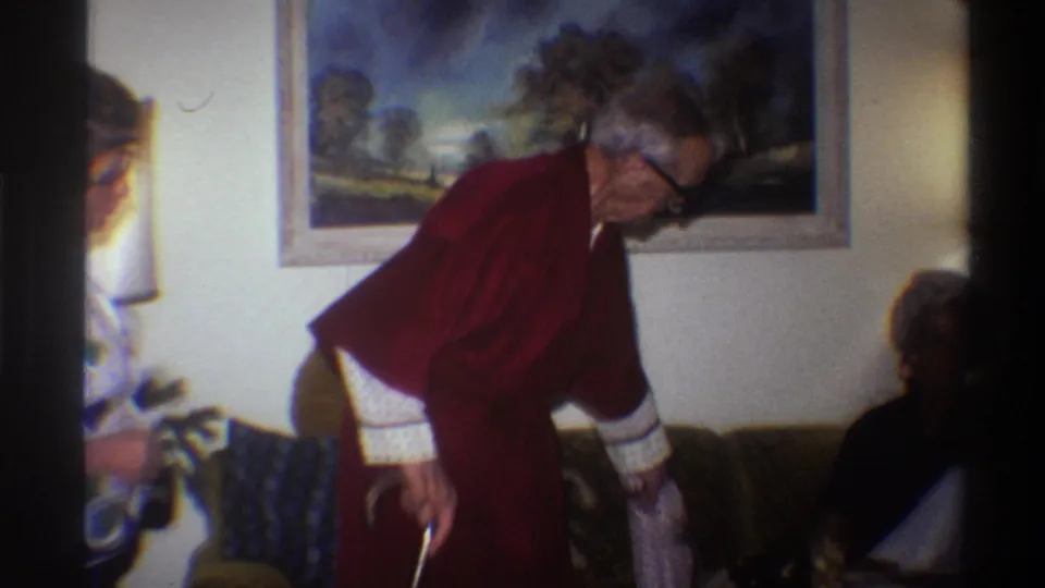 Mid-shot from a 1975 8mm home movie: elderly man in dark red robe with cane bending toward couch; partial silhouette of a second figure visible at right edge; framed landscape painting on wall.