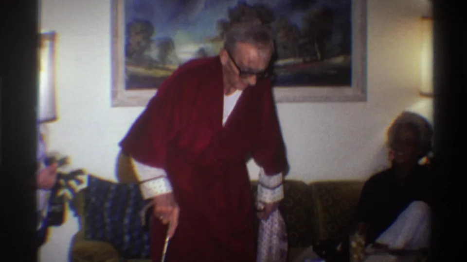 1975 8mm home movie frame: elderly man in red robe with glasses and white-tipped cane bends toward a seated figure on an olive-green couch; dark landscape painting visible on wall above.