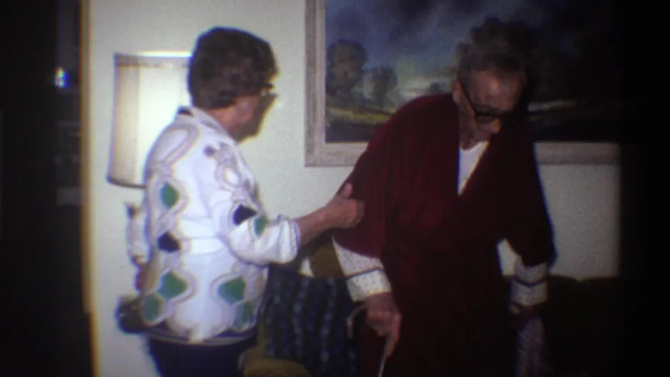 Closing frame of a 1975 8mm home movie: a woman in a bold geometric-print blouse steadies an elderly man in a dark red robe; floor lamp glowing at left, framed painting on wall behind.