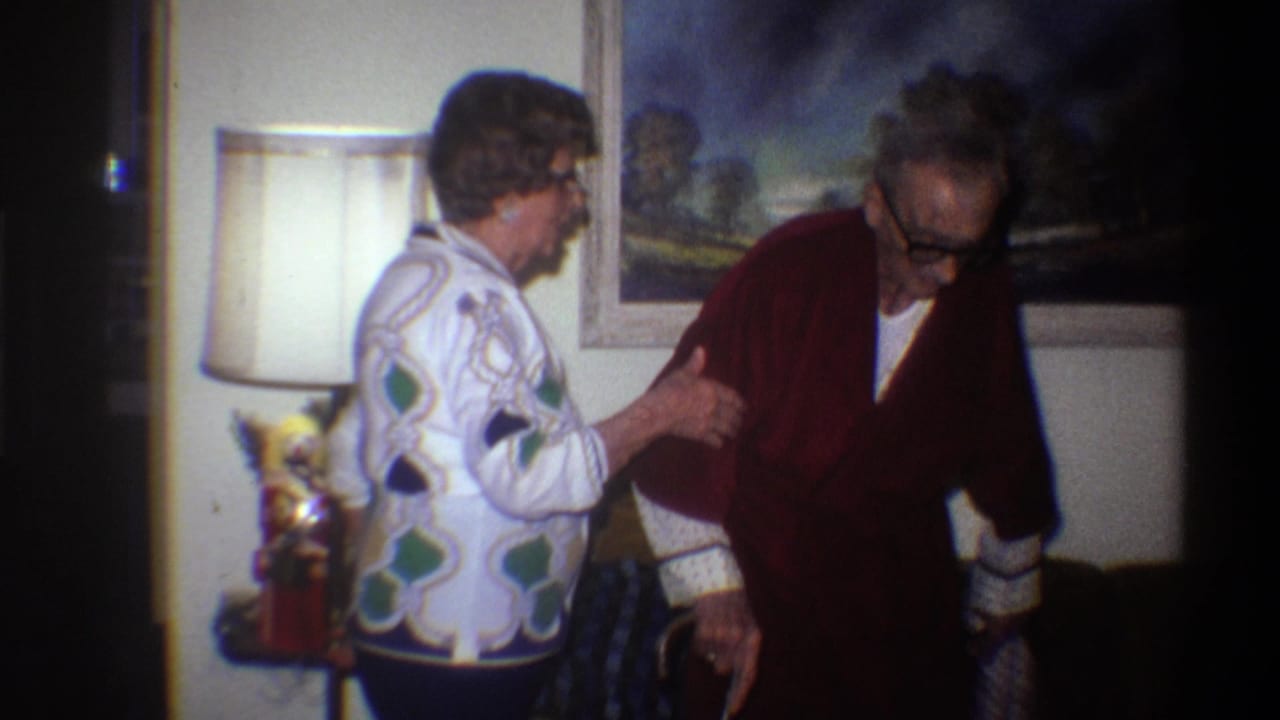 Elderly woman in a bold geometric jacket supports an older man in a dark red robe inside a 1975 Gainesville, Florida home interior