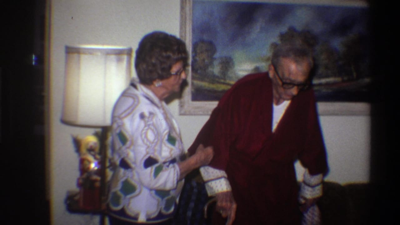 Older man in glasses and dark robe leans toward a woman in a geometric patterned jacket; lamp and framed painting visible behind them