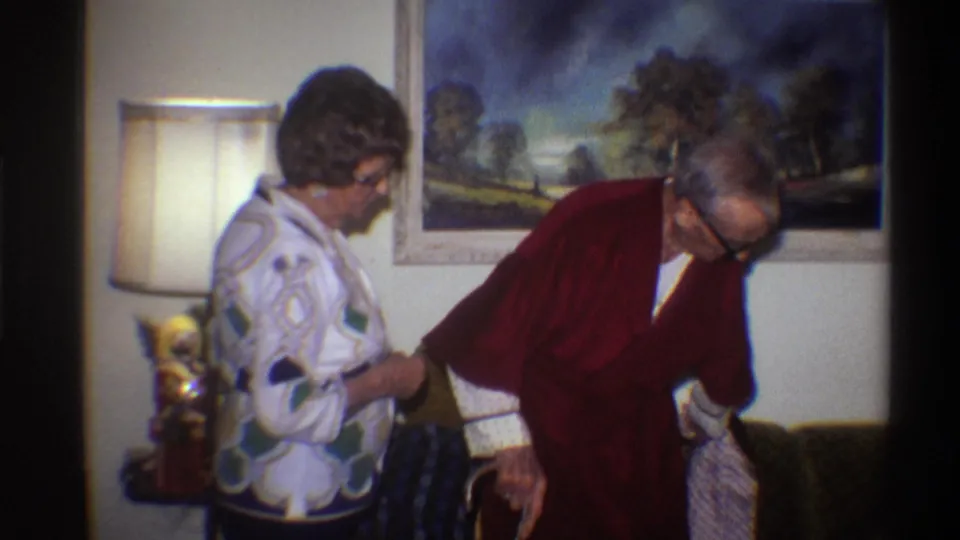 An elderly man in a dark red robe bends forward with apparent difficulty as a woman supports him; lamp and landscape painting visible in a 1970s home interior