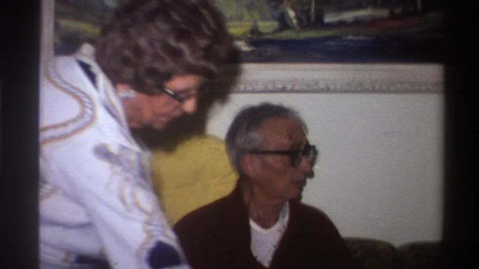 Close-up of an elderly man with thick-framed glasses looking down, with a woman in a geometric-patterned jacket beside him; yellow fabric and partial landscape painting visible