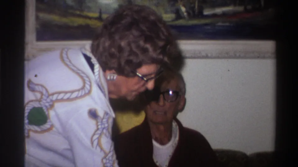 Two women in a 1975 Gainesville Florida home movie; the standing woman's embroidered white sweater and voluminous curly hair place the clip in mid-1970s domestic Americana.