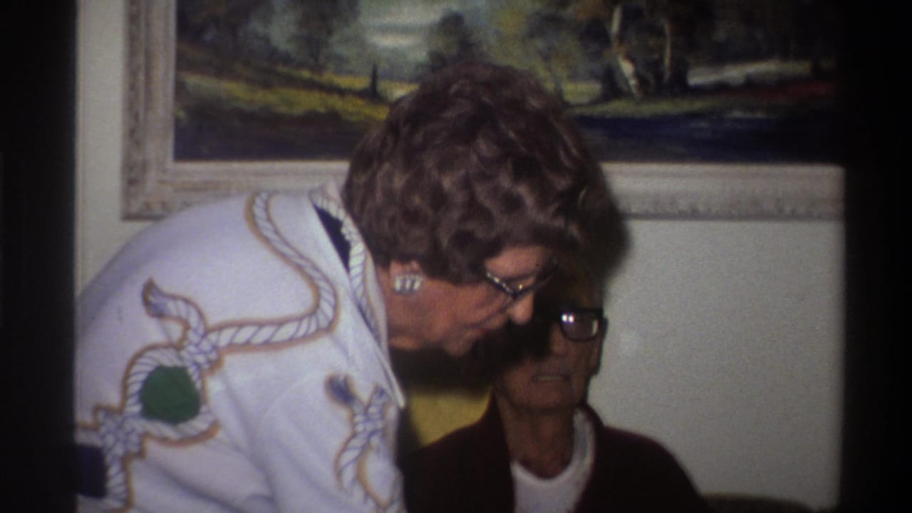 Closer view of two women; the standing woman in a white rope-motif sweater leans toward a seated older woman in a dark cardigan, both wearing glasses.