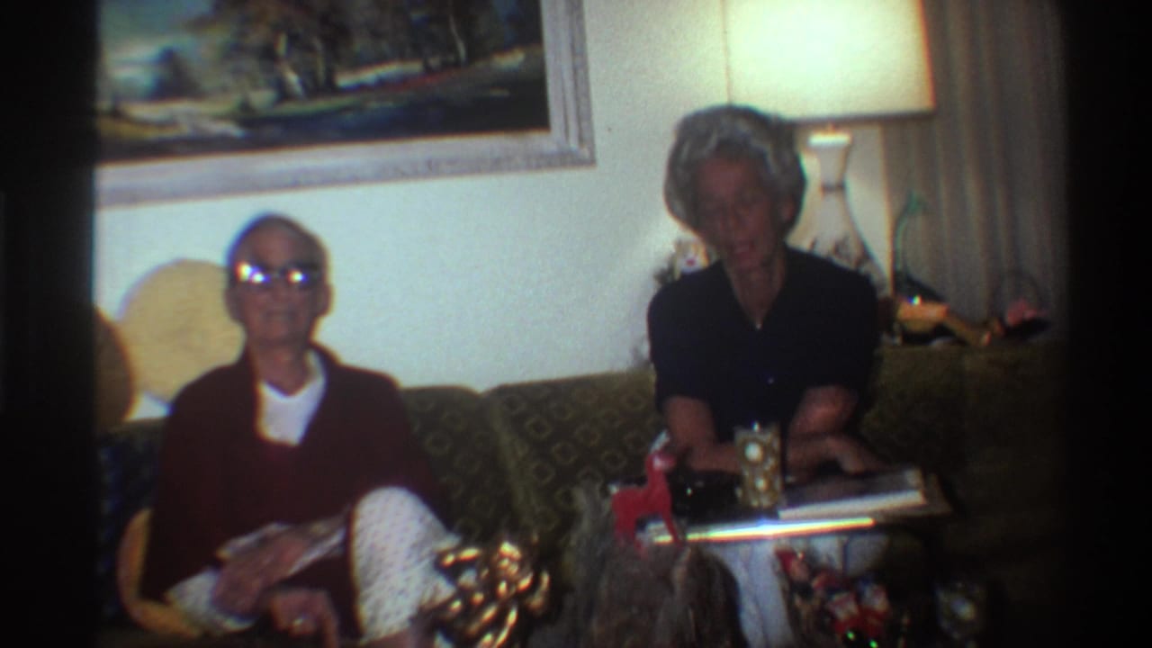1975 Gainesville, Florida — elderly man in glasses and dark red sweater and older woman with styled gray hair on a patterned couch, wrapped Christmas gift visible in foreground, 8mm home movie.
